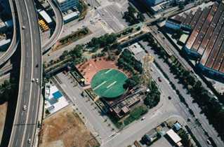 左側に幹線道路が走り、中央に緑色の円形の敷地と右手に黄色のタワーがある近隣一帯を上空から撮影した航空写真