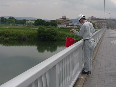 緑や家屋の手前に川があり、橋の上の白い手すりから乗り出すようにして作業服を着た男性が紐付きの赤いバケツで水をくみ上げて採水する様子の写真
