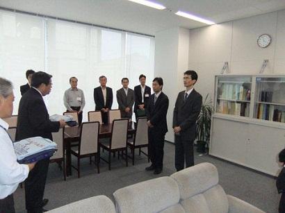 執務室で、二人の職員が、作業服を持った企業長と向かい合うように立っている写真