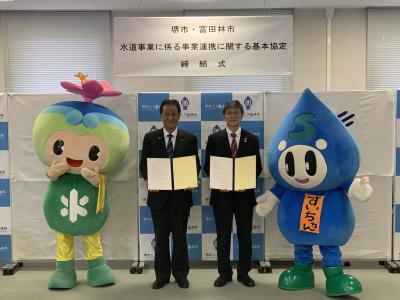 キャラクターのとっぴーとすいちゃんの間にスーツ姿の男性が2名それぞれ協定書を持って記念撮影をしている写真
