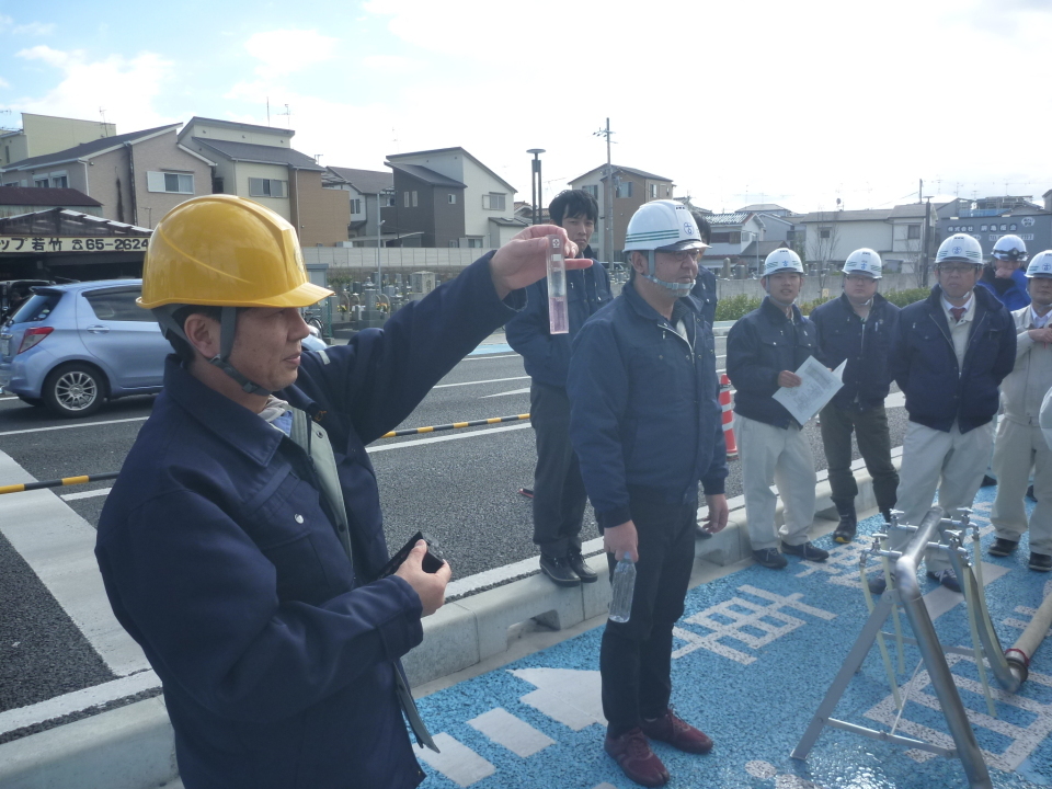 黄色のヘルメット姿の男性が水質確認している様子を周りの参加者にみせている写真